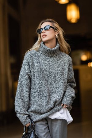Téléchargez les photos : Femme blonde dans des lunettes de soleil élégantes et pull gris debout avec sac à main noir à New York - en image libre de droit