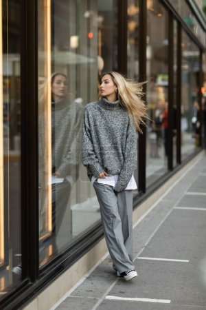 junge und stilvolle Frau im grauen Outfit, die in der Nähe von Schaufenstern in New York steht 