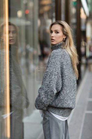 Blonde Frau im grauen Winteroutfit steht in New York vor einer Glasvitrine 