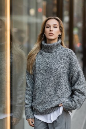 junge Frau im Winteroutfit steht mit der Hand in der Tasche in der Nähe von Schaufenstern in New York 