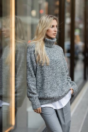 Blondine im Winteroutfit steht mit Hand in Tasche in der Nähe eines Schaufensters in New York 