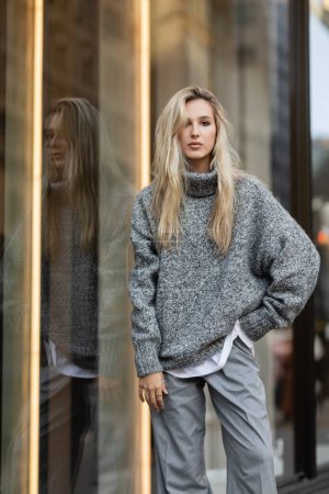 blonde young woman in winter outfit posing with hand in pocket near window display in New York 