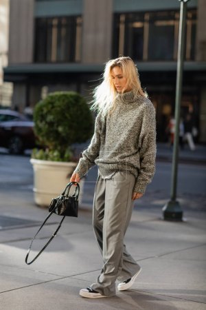 full length of stylish young woman with blonde hair holding handbag while walking in New York city in winter 