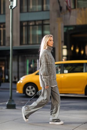 Stilvolle junge Frau mit blonden Haaren, die in der Nähe des gelben Taxis in New York spaziert 