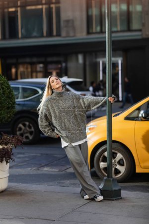 Stilvolle junge Frau im trendigen Winteroutfit posiert in New York in der Nähe von Straßenstange und gelbem Taxi 