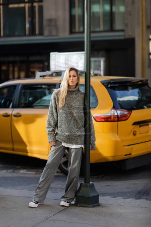 pełna długość młodej kobiety w szarym stroju zimowym pozowanie w pobliżu bieguna ulicy i żółtej taksówki w Nowym Jorku 
