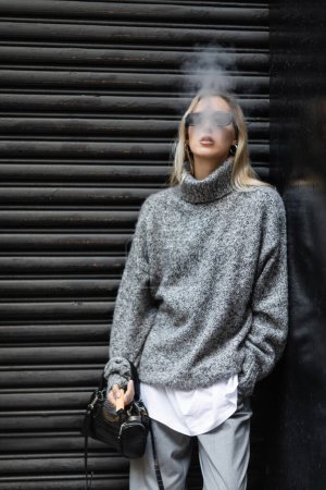 Blondine im Winterpullover und Sonnenbrille raucht E-Zigarette, während sie mit Handtasche auf der Straße steht 