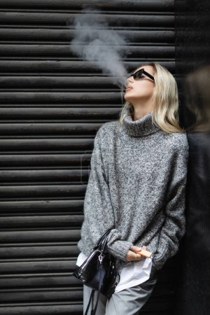 young woman in winter sweater and sunglasses smoking e-cigarette while standing with handbag on urban street 