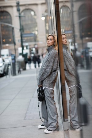 full length of blonde woman in winter sweater standing with handbag on urban street in New York 