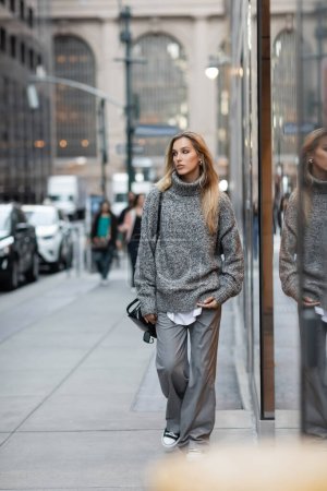 full length of blonde woman in winter sweater holding sunglasses and handbag while walking on urban street in New York 