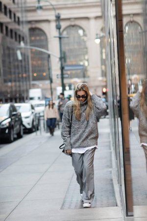 Frau in Winterpullover und Sonnenbrille läuft mit Handtasche auf urbaner Straße in New York 