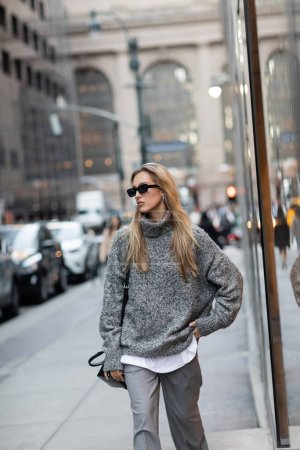 Hübsche Frau in Winterpullover und Sonnenbrille, die mit der Hand auf der Hüfte auf einer urbanen Straße in New York steht 