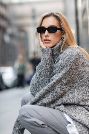 Blonde Frau im grauen Winterpullover und trendiger Sonnenbrille auf der Straße in New York City 