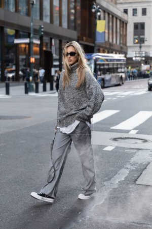 Photo for Full length of blonde woman in stylish knitted sweater and sunglasses walking on urban street in New York - Royalty Free Image