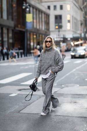 Téléchargez les photos : Femme blonde pleine longueur en pull tricoté et lunettes de soleil marchant la main dans la poche sur la rue urbaine de New York - en image libre de droit