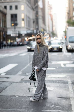 femme blonde pleine longueur en pull tricoté et lunettes de soleil marchant avec sac à main à la mode dans la rue urbaine de New York 