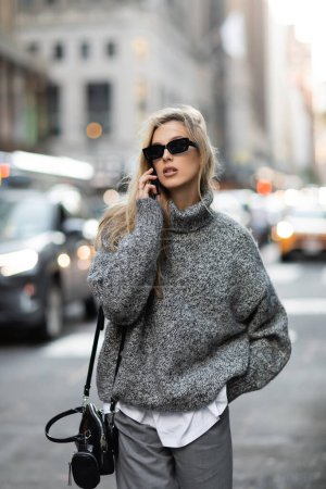 Stylische blonde Frau in Winterpullover und Sonnenbrille spricht in New York City auf dem Smartphone 