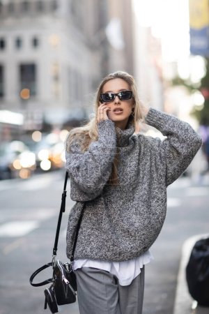 Blonde Frau in Winterpullover und Sonnenbrille spricht in New York City mit Smartphone 