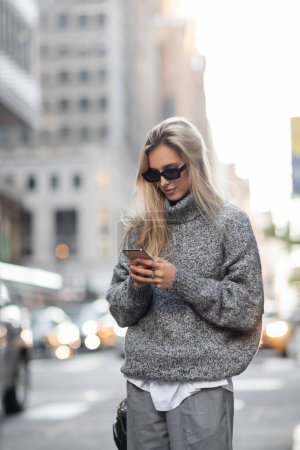blonde woman in knitted sweater and sunglasses using smartphone on urban street in New York city 