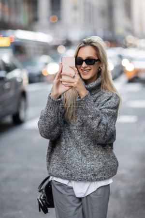 mujer joven feliz en suéter de invierno y gafas de sol tomando fotos en el teléfono inteligente en la ciudad de Nueva York 