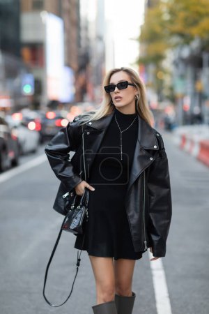 stylische Frau in Lederjacke und schwarzem Kleid, die mit der Hand an der Hüfte auf der Straße von New York steht 