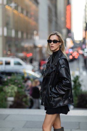 stylische Frau in Lederjacke und schwarzem Kleid mit Hand in Tasche in New York 