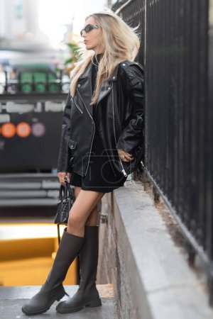 longitud completa de la mujer con estilo en chaqueta de cuero negro y gafas de sol de pie cerca de la entrada del metro en Nueva York 