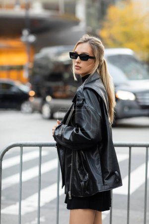 stylish woman in black leather jacket and sunglasses standing near metal barrier in New York 