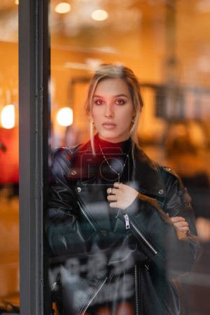 Foto de Mujer joven rubia en chaqueta de cuero negro mirando a la cámara a través de la ventana de cristal en Nueva York - Imagen libre de derechos