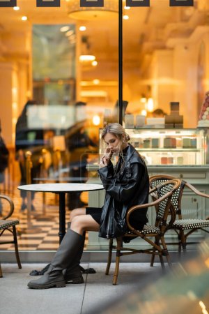 full length of blonde woman in black leather jacket sitting near round bistro table in outdoor cafe in New York 