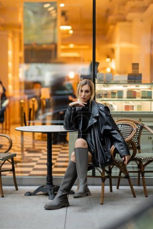 Blonde Frau in Lederjacke in voller Länge sitzt in der Nähe eines runden Bistrotisches in einem Café in New York 
