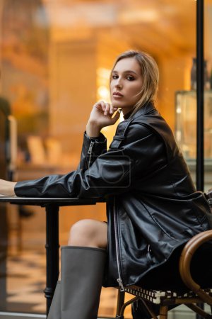 Eine junge Frau in schwarzer Lederjacke sitzt in voller Länge in der Nähe eines runden Bistrotisches in einem Café in New York 