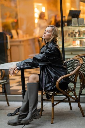 Stilvolle Frau in schwarzer Lederjacke und Stiefeln, die in der Nähe eines Bistrotisches in einem Café in New York sitzt 