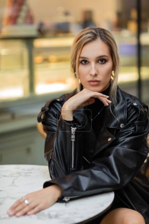 Photo for Young blonde woman in black leather jacket sitting near bistro table in outdoor cafe in New York - Royalty Free Image