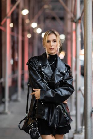 young blonde woman in black leather jacket standing on street in New York city at evening time