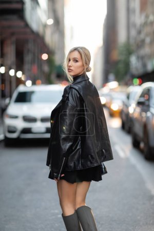 Blonde Frau in Lederjacke und schwarzem Kleid steht neben verschwommenen Autos auf der Straße in New York City 