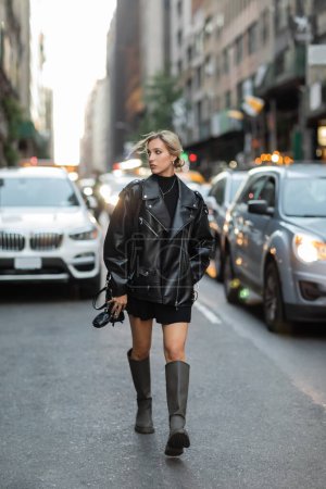 Frau in stylischer Lederjacke und schwarzem Kleid läuft in der Nähe verschwommener Autos auf der Straße in New York