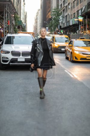 Blonde Frau in schwarzer Lederjacke und Kleid, die in New York in der Nähe von Autos und gelben Taxis spaziert 
