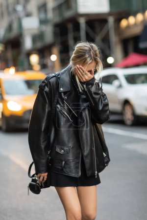Blondine in schwarzer Lederjacke und Kleid verdeckt Gesicht mit Hand auf Straße in New York 