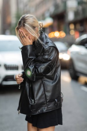 Téléchargez les photos : Femme blonde en cuir noir veste couvrant visage avec la main sur la rue à New York - en image libre de droit