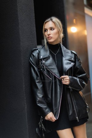 blonde woman in stylish leather jacket standing with handbag and looking away on urban street 