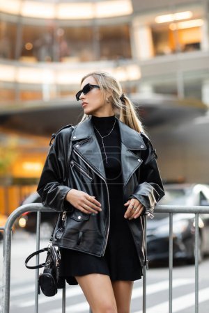 Stylische Frau in Lederjacke und Kleid steht mit Handtasche in der Nähe von Metallbarriere in New York 