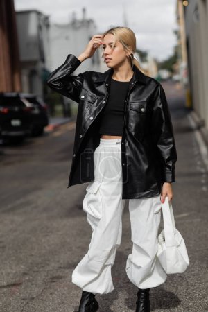 junge blonde Frau in stylischem Outfit hält Handtasche, während sie auf der Straße in Miami steht 