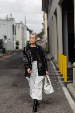 Junge Frau in stylischem Outfit hält Handtasche, während sie auf der Straße in Miami spaziert 