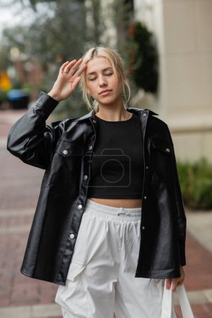 young woman in leather shirt jacket standing with handbag and adjusting blonde hair 