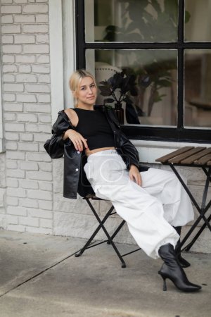 Téléchargez les photos : Pleine longueur de femme blonde en veste chemise en cuir et pantalon cargo assis dans un café extérieur dans la rue à Miami - en image libre de droit