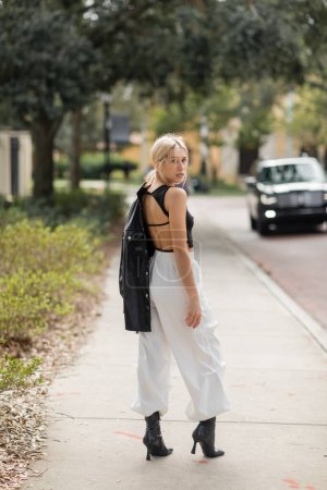 Blonde Frau in weißen Cargohosen und Stiefeln steht mit schwarzer Lederjacke auf der Straße in Miami  