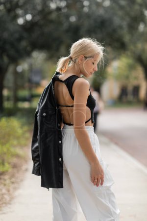 Téléchargez les photos : Vue latérale de la femme blonde en pantalon cargo blanc debout avec veste en cuir noir sur la rue à Miami - en image libre de droit