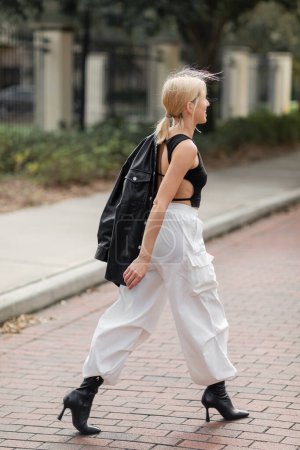 Téléchargez les photos : Pleine longueur de femme gaie en pantalon cargo blanc et bottes marchant avec veste en cuir noir sur la rue à Miami - en image libre de droit
