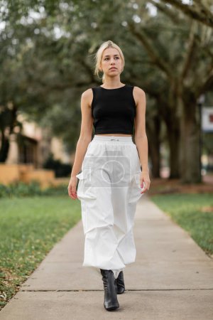 Téléchargez les photos : Pleine longueur de jeune femme en pantalon cargo blanc et débardeur noir marchant sur la rue verte à Miami - en image libre de droit
